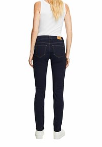 Žena s dlouhými blond vlasy má na sobě tmavě modré skinny džíny, bílé bezrukávové tričko a bílé platformové tenisky, stojí zády.