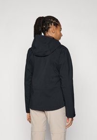 Black hooded jacket with a smooth texture, featuring a cinched waist and zippered pockets, viewed from the back, worn with light-colored pants.