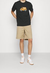 Hombre vestido con una camiseta negra con un gráfico de furgoneta naranja, pantalones cortos beige, calcetines blancos y zapatillas blancas con acentos naranjas, de pie con las manos detrás de la espalda.