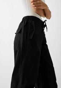 Svarta cargobyxor med elastisk midja, snörning och flera fickor. Tyget har en texturerad yta och avslappnad passform.