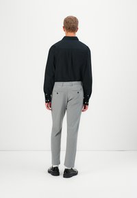 Homme portant une chemise noire à manches longues, un pantalon de costume gris clair, des chaussettes blanches et des chaussures de cuir noires, se tenant face à un mur blanc.