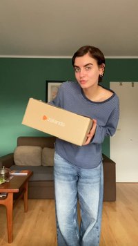 Caixa de cartão da Zalando com o logótipo laranja, segurada por uma pessoa vestindo uma camisa de mangas longas azul às riscas e jeans azuis claros, num ambiente de sala de estar.