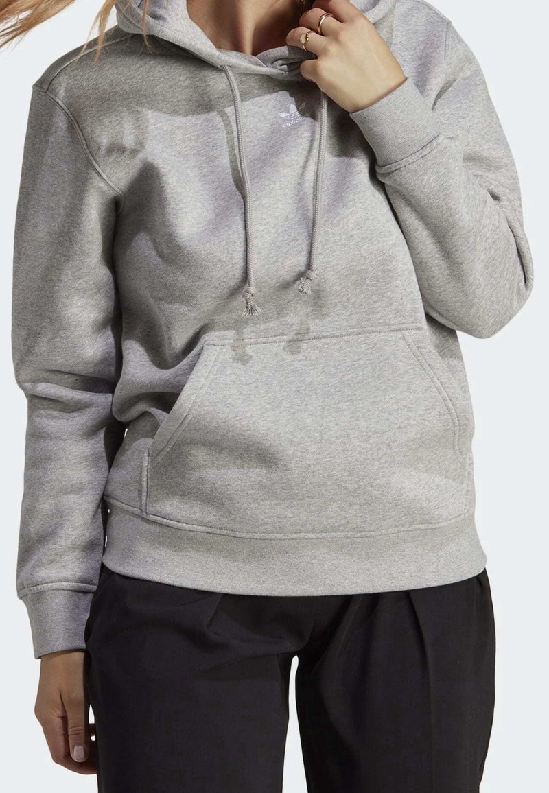 Grå huva sweatshirt gjord av mjukt tyg, med en framficka, dragsnoddar och ribbade ärmslut, visas med svarta byxor.