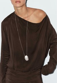 Brązowa, luźno zwisająca bluzka z długim rękawem i fasonem odsłaniającym ramiona; srebrny owalny wisiorek zawieszony na cienkim łańcuszku. Gładka, miękka tekstura.