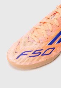 Primo piano della punta di una scarpa da calcio indoor arancione con il testo "F50" e "SPRINTGRID" in blu e lacci pesca su uno sfondo chiaro.