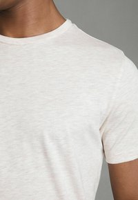 Tricou scurt din bumbac de culoare bej deschis, confecționat dintr-un material moale cu o textură subtilă, având un guler clasic și detalii de coasere curate.