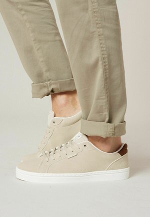 Personne portant des baskets basses beige et un pantalon kaki retroussé, debout sur une surface unie de couleur claire.