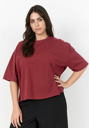 Donna con lunghi capelli scuri che indossa una camicia rossa a maniche corte e pantaloni neri, in piedi con una mano sui fianchi contro uno sfondo bianco.