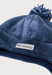 Modrá fleecová zimná čiapka s pom-pomom na vrchu, s vyšitým logom Columbia na prednej strane. Mäkká textúra a zaoblený tvar.