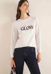 Světle béžová mikina s dlouhým rukávem, na které je tmavý nápis "Mountain GLOW" a "REACH HEIGHTS." Kombinováno s tmavě modrými džíny se bílým prošitím.