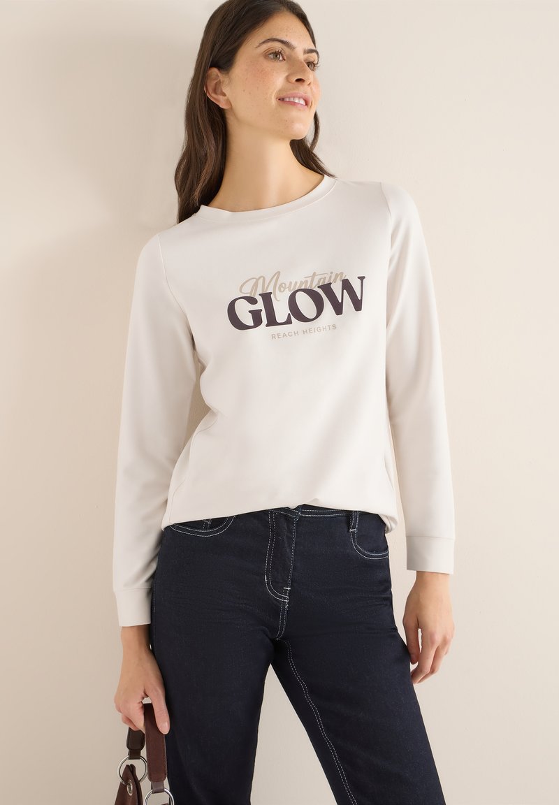 Světle béžová mikina s dlouhým rukávem, na které je tmavý nápis "Mountain GLOW" a "REACH HEIGHTS." Kombinováno s tmavě modrými džíny se bílým prošitím.