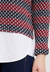 Pull en maille aux motifs rouge, bleu marine et blanc ; design court avec poignets côtelés. Superposé sur une chemise blanche et un jean bleu au niveau de l'ourlet.