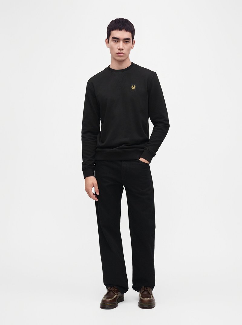 Jeune homme portant un sweat-shirt noir avec un petit logo sur la poitrine, un pantalon noir et des chaussures marron, debout avec une main dans la poche.