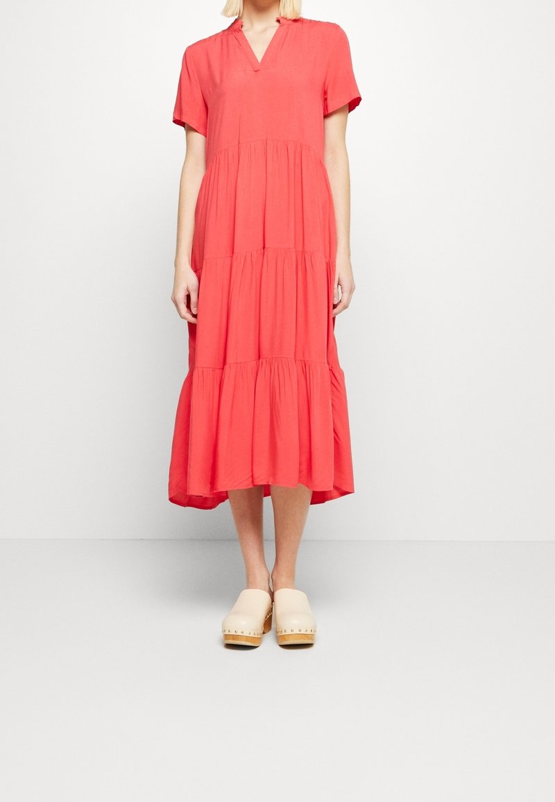 Femme portant une robe midi corail à volants avec manches courtes et des sabots en bois beige, debout devant un fond blanc uni.
