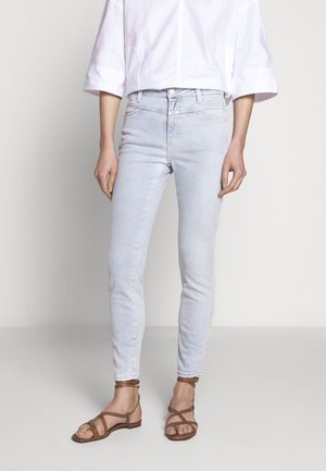 Kapealahkeiset vaaleansiniset farkut, yhdistettyinä ruskeisiin remmikkäisiin tasapohjaisiin sandaaleihin ja löysään valkoiseen lyhytpaitaiseen topiin seisovan henkilön päällä.