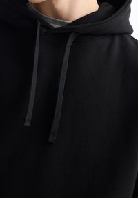 Svart hoodie i mjukt tyg med dragsko i huvan, platta sömmar och en slät yta. Lager ovanpå ett grått plagg.