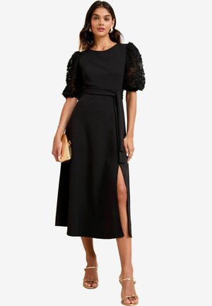 Zwarte jurk met ingewikkelde kant pofmouwen, getailleerde lijfje, tailleband en hoge split. Draagt met gouden sandaaltjes en een kleine gouden clutch.