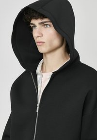 Sweat à capuche zippé noir en tissu lisse et texturé. Présente une grande capuche et des poches latérales. Porté sur une chemise rose clair.