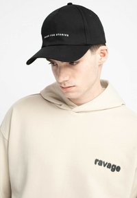 Młody mężczyzna w czapce z daszkiem w kolorze czarnym z napisem "MADE FOR STORIES" oraz beżowym hoodie z napisem "ravage" na klatce piersiowej, patrzy w dół.