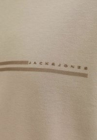 Un textile beige à texture lisse présente un logo en relief discret "JACK&JONES" et deux lignes horizontales dans une teinte légèrement plus foncée.