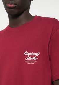 Sarkans kokvilnas T-krekls ar apaļu kakla izgriezumu un īsām piedurknēm. Uz kreisā krūšu apgabala ir balts teksta logs. Gluda tekstūra, ikdienas piegriezums.