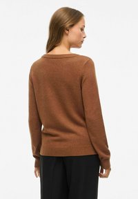 Brązowy sweter robiony na drutach z okrągłym dekoltem, ściągaczami przy mankietach i na dolnej krawędzi, wykonany z miękkiego materiału, pokazany z tyłu na jasnym tle.