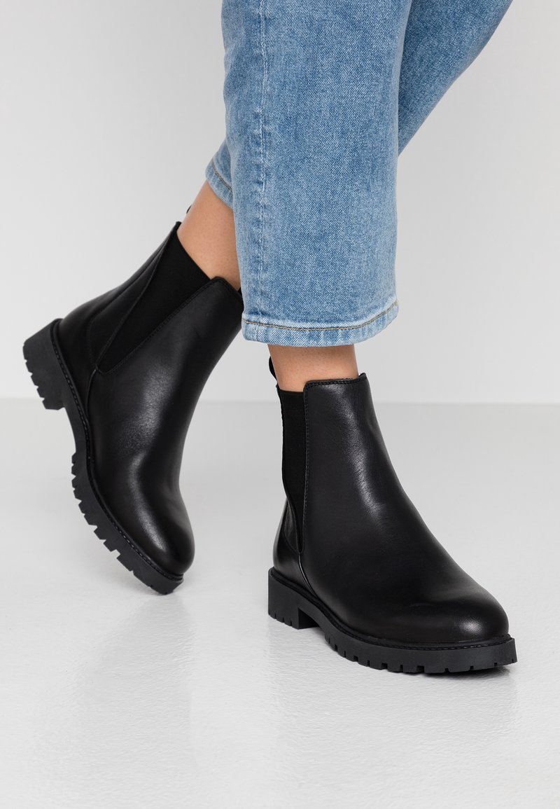 Personne portant des bottines en cuir noir à semelles épaisses et un jean court bleu clair, debout sur un sol blanc.