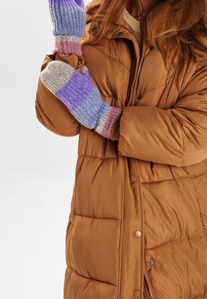 Personne portant une veste matelassée marron avec des moufles multicolores en tricot, tenant un bras croisé devant son corps sur un fond uni.
