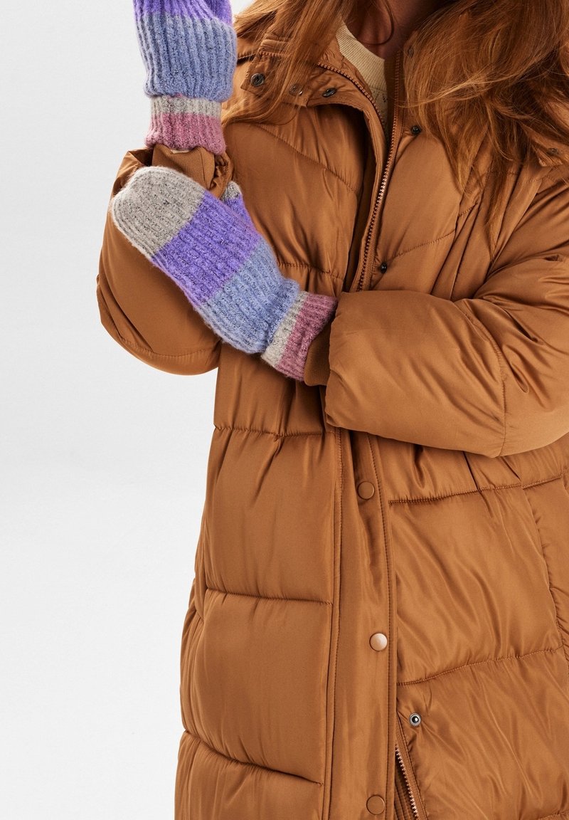 Personne portant une veste matelassée marron avec des moufles multicolores en tricot, tenant un bras croisé devant son corps sur un fond uni.