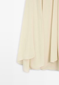 Světle béžová tkanina s hladkým povrchem, která má měkké záhyby a nepravidelný lem. Minimalistický design bez viditelných zapínání.