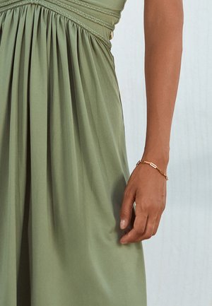 Bras de femme portant un délicat bracelet en or, à côté d'une robe plissée vert olive avec un détail de taille froncée.