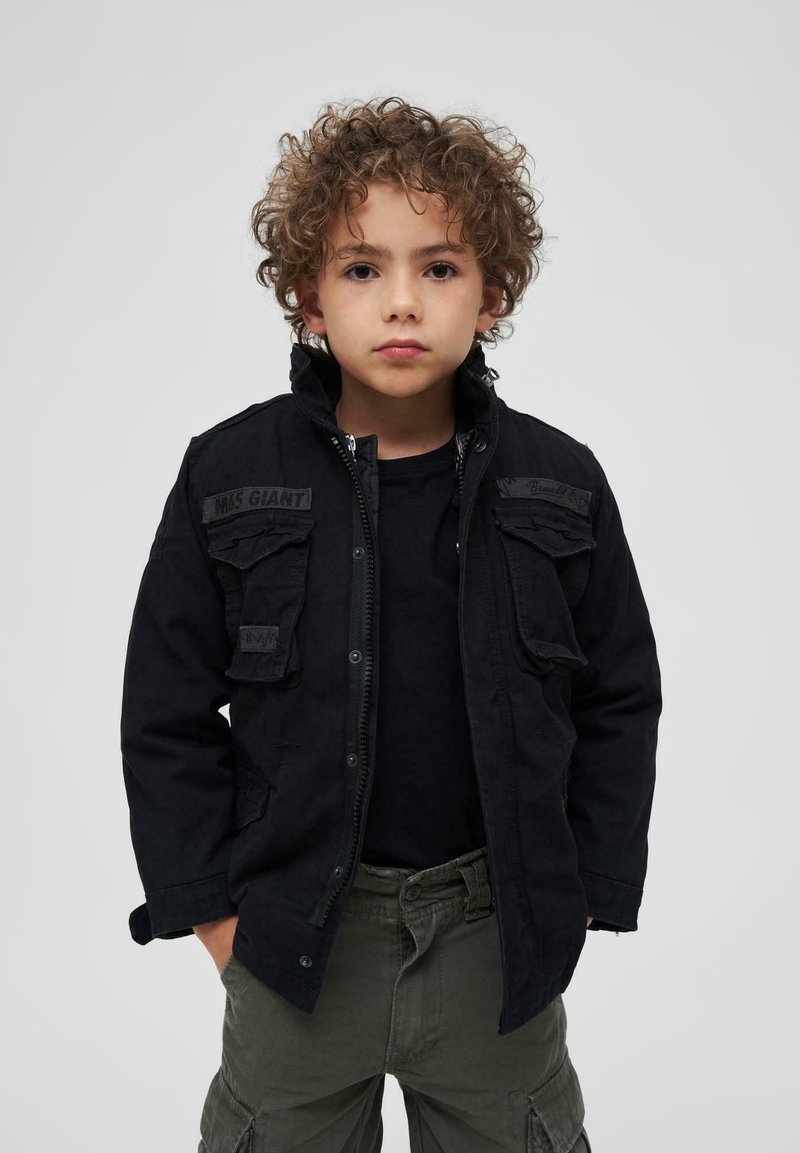 Juodas kariškas stiliaus švarkas su priekinėmis kišenėmis, užtrauktuku ir tvirtu audiniu. Dėvimas ant juodos palaidinės ir žalių kargo kelnių.