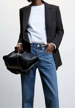 Personne portant un blazer noir, une chemise blanche, un jean bleu, tenant un grand sac à main en cuir noir sur le côté avec une main dans la poche.