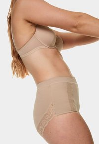 Béžová podprsenka a souprava kalhotek s vysokým pasem. Podprsenka má hladkou tkaninu a krajkové detaily; kalhotky mají krajkové ozdoby po stranách.