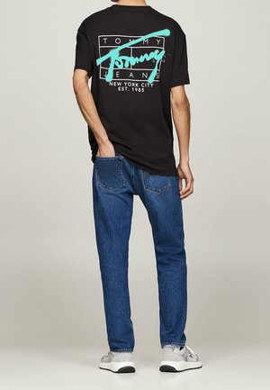 Male model wearing black graphic Tommy Jeans t-shirt, blue jeans, and gray sneakers, standing with one hand in back pocket facing away.