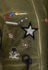 Veste vert olive ornée de cristaux décoratifs, d'un emblème de crâne et de patchs étoilés avec des accents noirs et dorés. Tissu texturé avec des détails brillants.