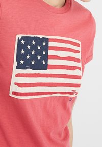 Červené bavlněné tričko s výraznou vyšitou grafikou americké vlajky v námořnické modři a bílé, s podrobnými hvězdami a pruhy.