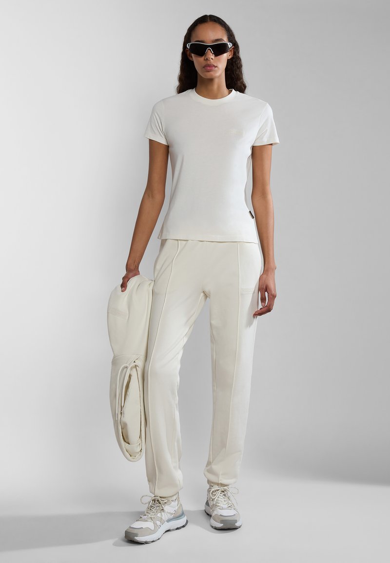 Weißes T-Shirt mit kurzen Ärmeln und passende schmale Jogginghosen, beide aus einem weichen Material. Zubehör umfasst sportliche weiße Sonnenbrillen und klobige Sneaker.