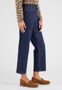 Jeans de mezclilla oscuros de tiro alto con un ajuste relajado, terminados con costuras en contraste y un cierre de botón, combinados con mocasines de ante color marrón.