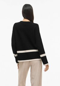 Melns džemperis ar apaļu kaklu, ar divām horizontālām krēmkrāsas svītrām apakšmalā un piedurknēs. Valkāts ar gaisvi bēšām biksēm.