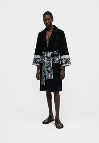 Homme portant une robe noire mi-longue ornée d'un galon argenté à motif et d'une ceinture, associée à des sandales noires, debout sur fond blanc.