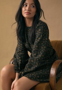 Sort kjole med gyldne trykte mønstre, lange ærmer og rund halsudskæring. Glat stof, knælængde, kombineret med en brun lænestol.