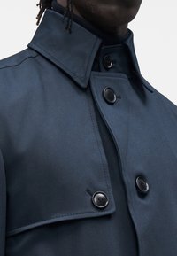 Palton trench bleumarin, cu guler structurat, partea frontală cu două rânduri de nasturi și nasturi negri. Materialul are o finisare netedă și un luciu subtil.