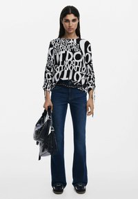 Maglietta a maniche lunghe bianco e nero con motivo, abbinata a jeans blu a campana. Modella che tiene due borse nere con accenti di borchie.