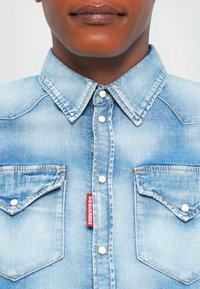 Detailný záber na osobu, ktorá má na sebe svetlomodrú denimovú košeľu na gombíky s špicatým golierom a dvoma náprsnými vreckami, z ktorých jedno má červený štítok DSQUARED2.