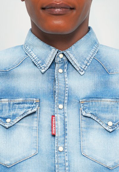 Close-up of a person wearing a light blue denim button-up shirt with pointed collar and two chest pockets, one with a red DSQUARED2 label.