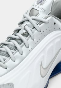 Close-up of gray Nike Shox sneaker showing laces, perforated side panel, textured tongue, and Nike swoosh on the white midsole.
