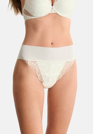 Femme portant de la lingerie en dentelle blanche avec une large ceinture et des détails de broderie florale à l'avant sur un fond uni.