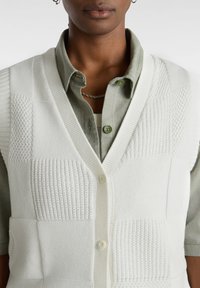 Hvid strikket vest med en blanding af teksturerede mønstre og V-hals design, med knaplukning, lagdelt over en olivengrøn skjorte.