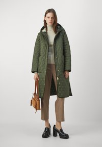 Cappotto lungo imbottito verde con cappuccio, accessoriato con hardware dorato, abbinato a un leggero maglione e pantaloni a scacchi. Borsa marrone e scarpe platform nere.